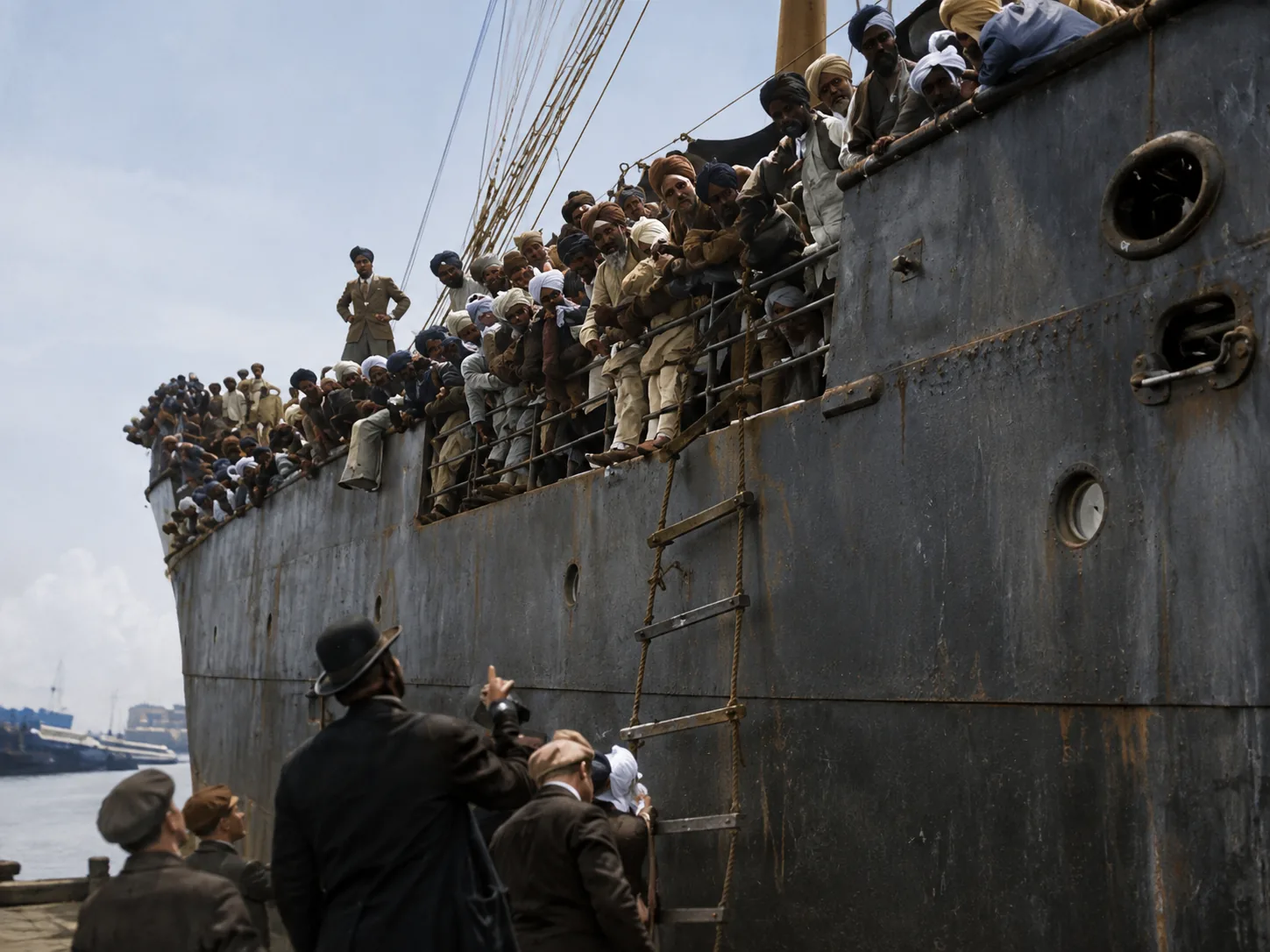 Illustration of passengers packed along a ship's rail high above a quay, watched from below by officials and a man with a camera.