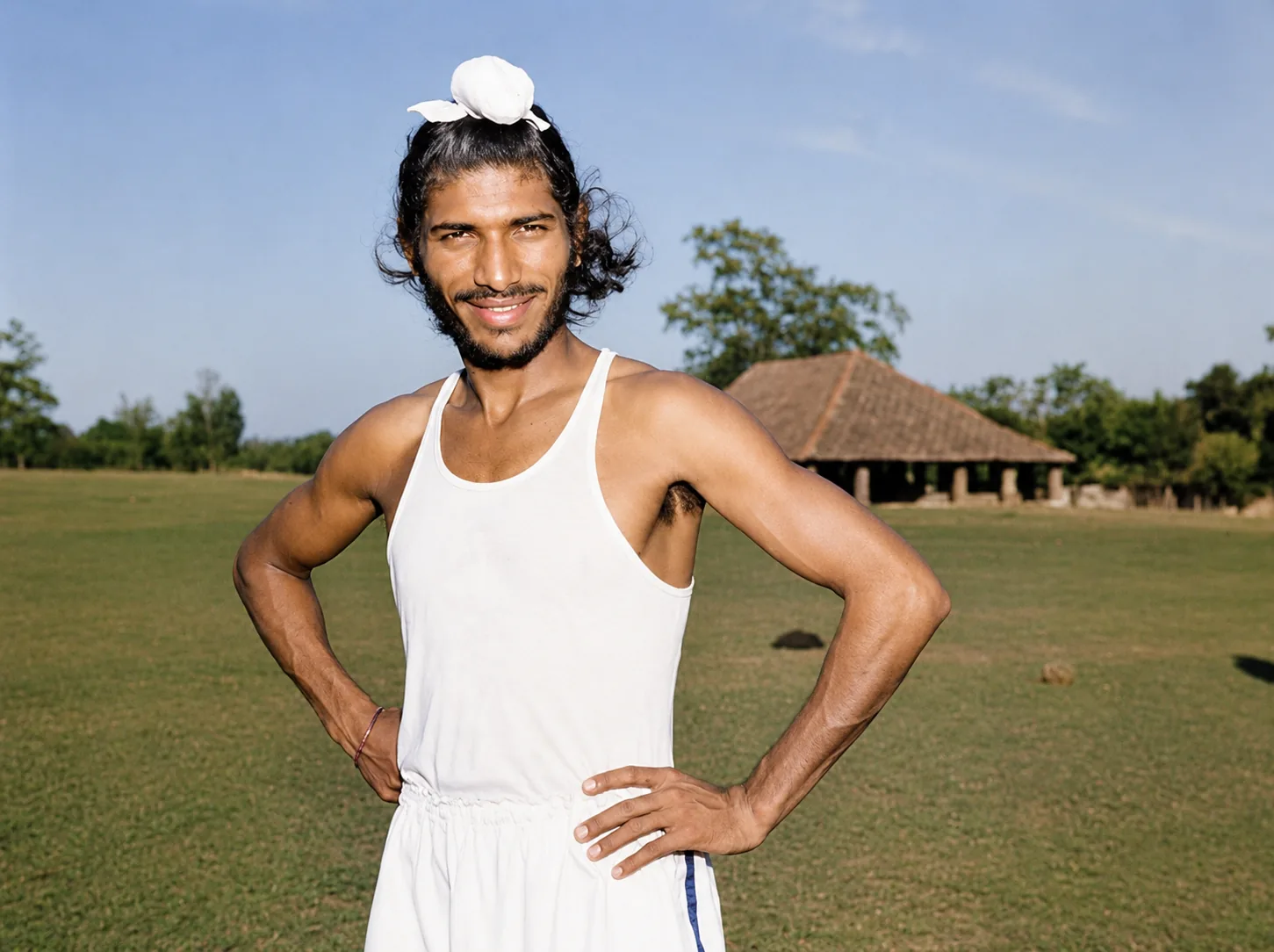 Illustration of Milkha Singh as a young man in a white running vest, hands on his hips, standing on grass in front of a tiled-roof building.